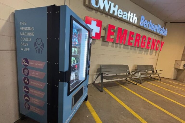 A vending machine with harm reduction supplies is placed near the entrance to an emergency room at UW Health, next to empty benches and emergency signs.