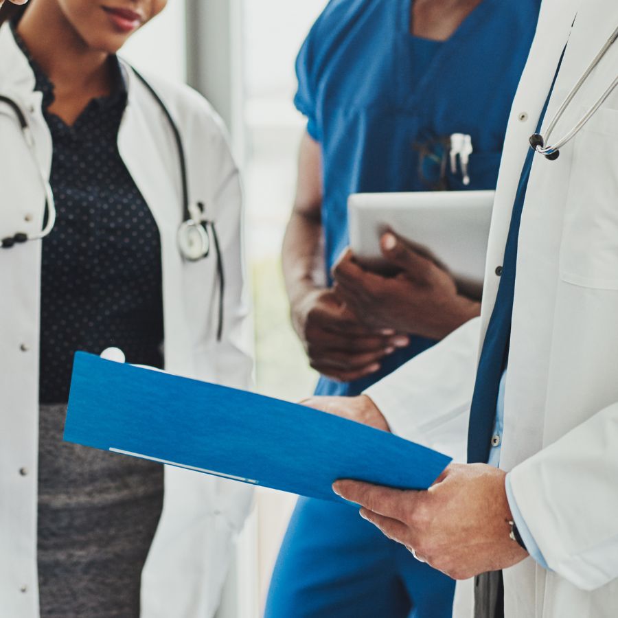 Three healthcare professionals, two in white coats and one in blue scrubs, stand together discussing documents held in a blue folder.