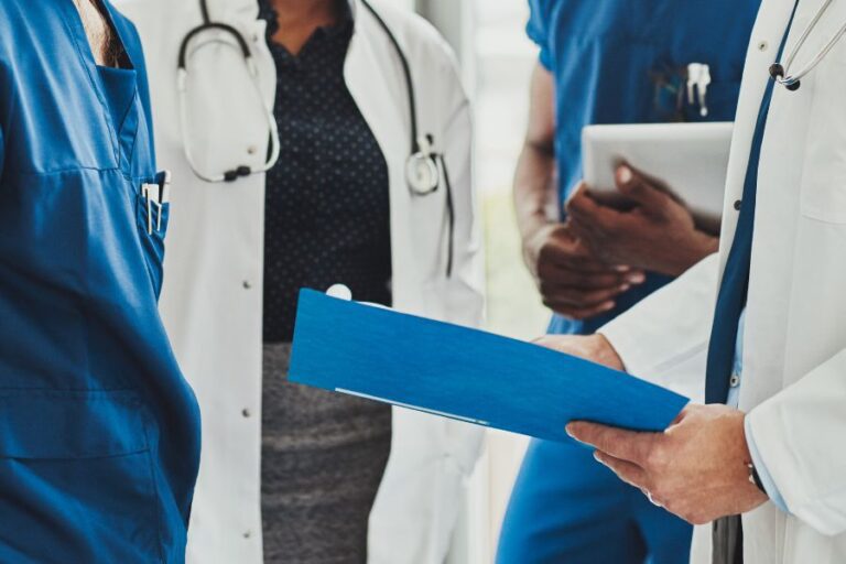 Three medical professionals in scrubs and lab coats stand together, one holding a blue clipboard, having a discussion in a clinical setting.