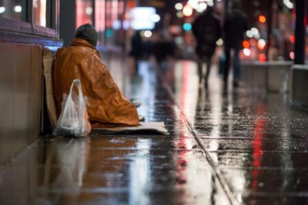 A person sits on cardboard under a blanket on a wet city sidewalk at night, with a plastic bag nearby and blurred pedestrians in the background.