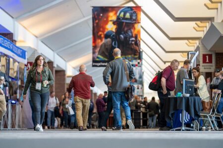 People walk and interact in a convention hallway with booths, banners, and a large firefighter poster in the background.