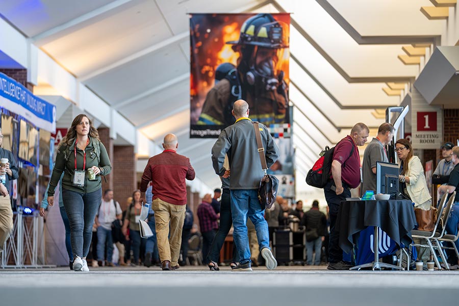 People walk through a convention hallway with booths on either side; a large firefighter banner hangs in the background.