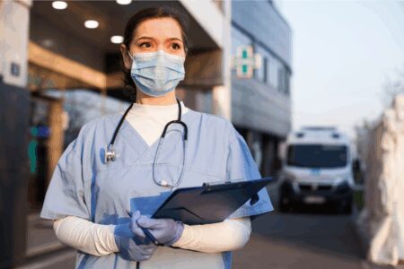 A healthcare worker wearing scrubs, gloves, and a mask stands outside a medical facility holding a clipboard, with an ambulance visible in the background.