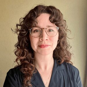 A woman with curly brown hair and glasses, wearing a dark collared shirt, stands in front of a beige wall and looks at the camera.