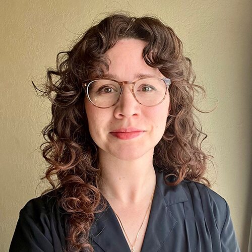 A woman with curly brown hair and glasses, wearing a dark collared shirt, stands in front of a beige wall and looks at the camera.