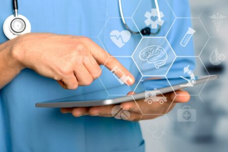 A person in medical scrubs uses a tablet with healthcare-related digital icons overlaid, including symbols for a heart, brain, and stethoscope.