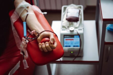 A person donates blood, squeezing a red stress ball, with blood flowing through a tube into a collection bag beside a medical machine.