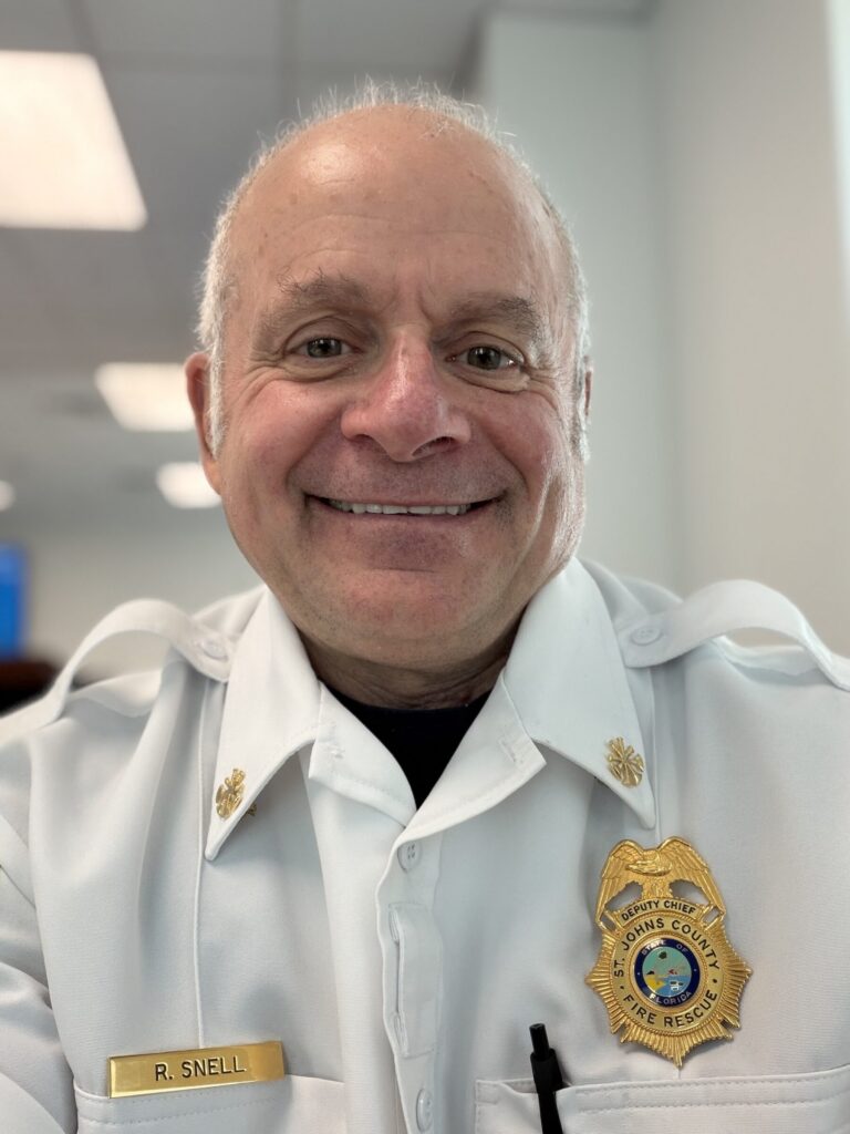 A man in a white fire rescue uniform with a badge and name tag reading "R. Snell" smiles at the camera in an indoor setting.