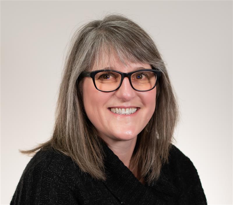 Woman with straight gray hair and glasses, wearing a black sweater, smiling at the camera against a plain light background.