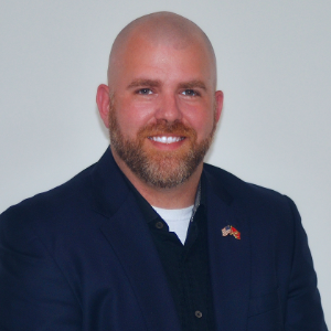 A man with a shaved head and beard, wearing a dark blazer and black shirt, smiles in front of a plain light background.