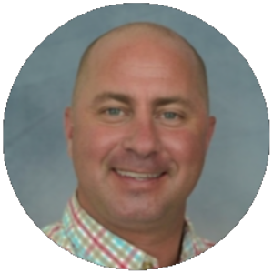 A man with a shaved head and light complexion, smiling, wearing a colorful plaid shirt, against a light blue-gray background.