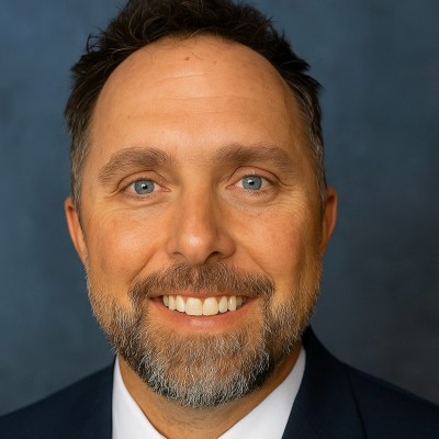 A man with short brown hair, blue eyes, and a trimmed beard wearing a dark suit jacket, white dress shirt, and smiling against a blue background.