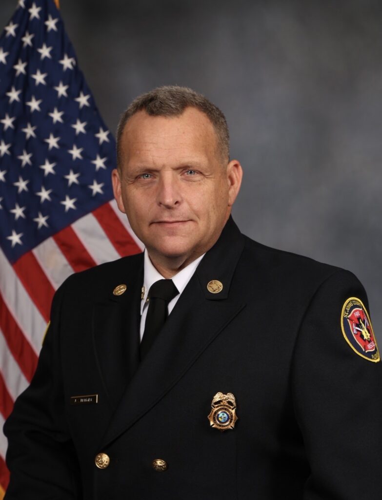 A man in a formal fire department uniform stands in front of an American flag, facing the camera with a neutral expression.