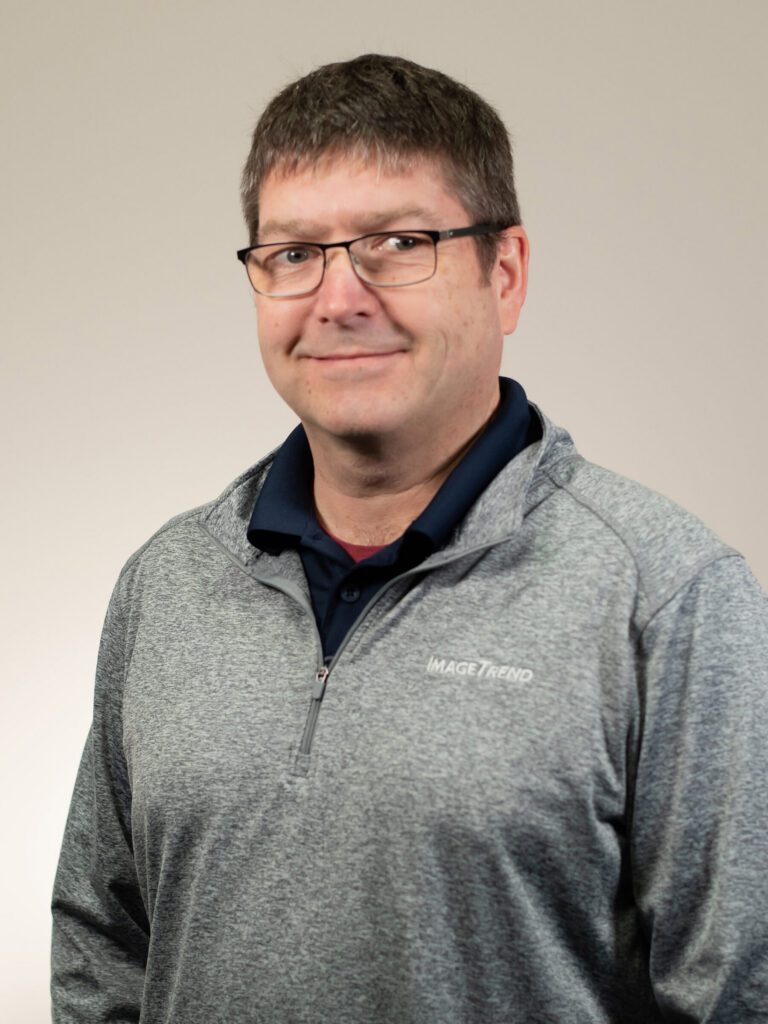 A man with short brown hair and glasses wearing a gray quarter-zip pullover with "IMAGETREND" logo stands against a plain light background, facing the camera and smiling slightly.
