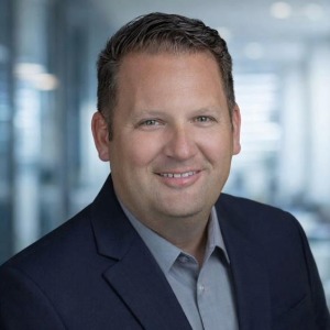 A man in a suit jacket and collared shirt smiles at the camera in an office setting with blurred background.