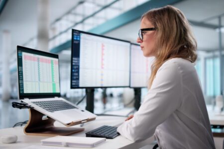 A woman wearing glasses works at a desk with a laptop on a stand and two large monitors displaying spreadsheets in an office setting.
