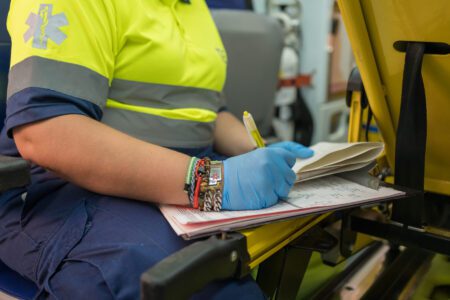 A paramedic wearing blue gloves and a yellow uniform writes notes on a clipboard inside an emergency vehicle.