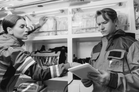 Two paramedics in uniform work inside an ambulance; one looks at a tablet while the other reaches for medical supplies on a shelf.