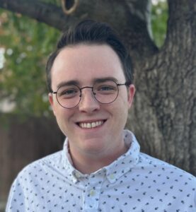 A person wearing glasses and a white shirt with small patterns stands outdoors in front of a tree, smiling at the camera.