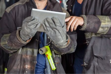 Two people in firefighting gear use a tablet. One wears gloves and holds the device, while the other points at the screen. A flashlight hangs from one person's jacket.