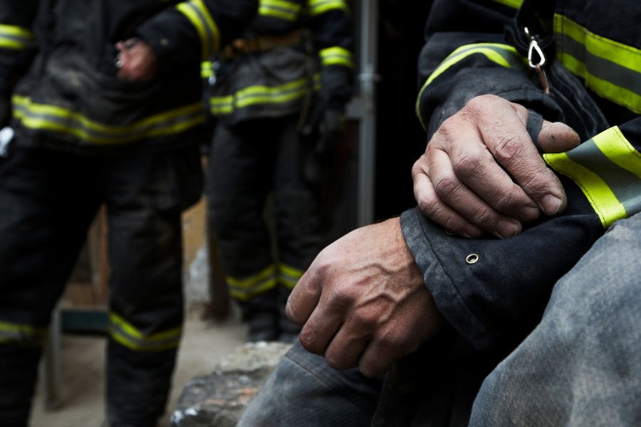 Close-up of two firefighters in protective uniforms with reflective stripes, one adjusting their sleeve, both with dirty hands, suggesting recent action or emergency response.