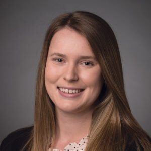 A woman with long brown hair smiles at the camera against a plain gray background. She is wearing a black jacket and a light-colored blouse.