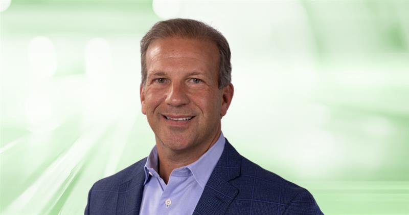 A man in a blue blazer and light blue shirt smiles at the camera against a blurred green and white background.