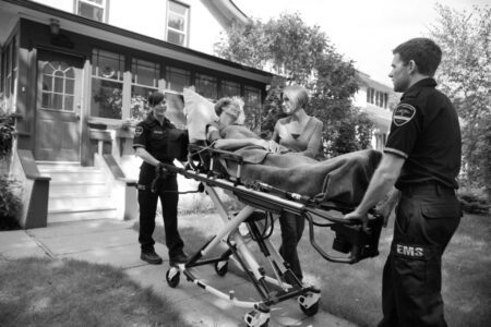 Two EMS workers wheel a person on a stretcher outside a house while another EMT and a woman stand nearby, talking to the patient.