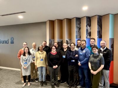 A group of fifteen people pose for a photo indoors in front of a modern wall with wood panels and images.