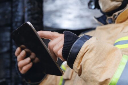 A person in firefighting gear uses a touchscreen tablet, focusing on the device with their finger.