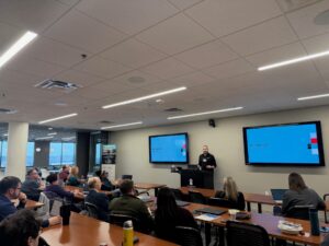 A person stands at a podium giving a presentation titled "Our Approach" to an audience in a modern conference room with two large screens displaying the slides.