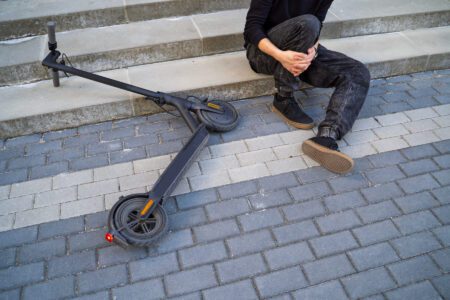 A person sits on steps holding their knee next to a fallen electric scooter on a paved sidewalk.