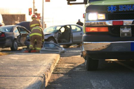 A firefighter attends to a car crash scene involving two vehicles, with an ambulance parked nearby on the street.