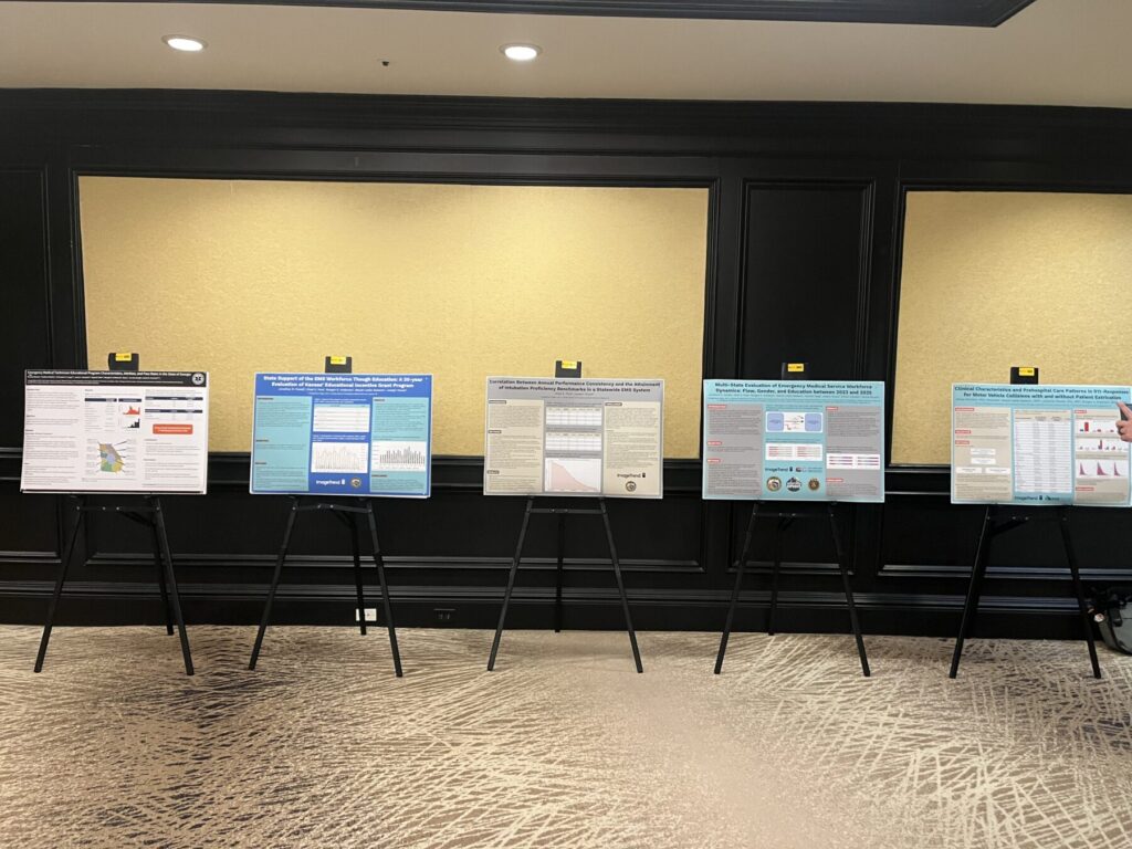 Five research posters displayed on easels in a carpeted conference room with black paneled walls and a yellow background.