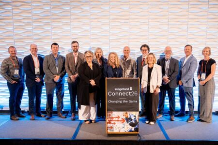 A group of thirteen people stands on a stage behind a podium labeled "ImageTrend Connect26: Changing the Game" against a textured white backdrop.