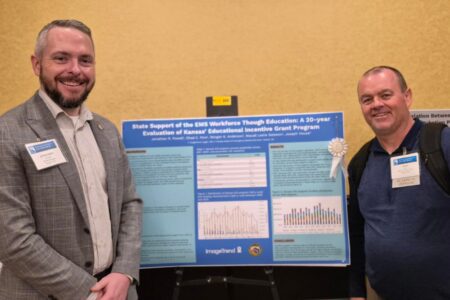 Two men stand beside a research poster titled "State Support of the EMS Workforce Through Education" at a conference, both wearing name tags and smiling at the camera.