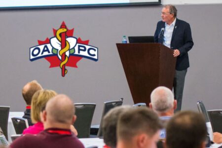A man stands at a podium giving a presentation to an audience; an OAPC logo with a maple leaf and caduceus is displayed on the wall.