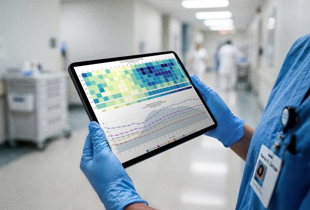 A healthcare worker wearing blue gloves holds a tablet displaying medical data and charts in a hospital corridor.
