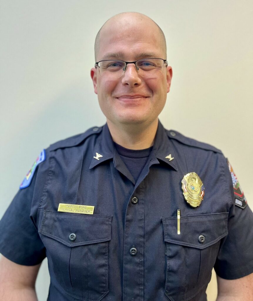 A person in a police uniform with badges and a nameplate stands facing the camera against a plain light background.