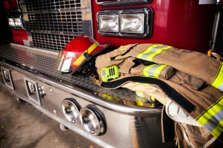 A firefighter’s helmet, jacket, and radio rest on the front bumper of a red fire truck inside a fire station.