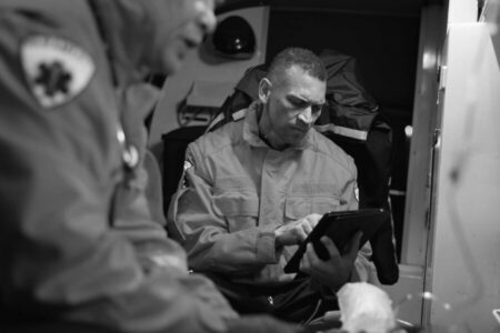 Two emergency responders in uniform are inside an ambulance; one is using a tablet while the other is partially visible in the foreground.