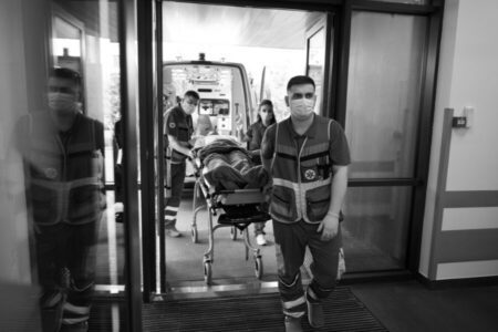 Paramedics wearing masks wheel a patient on a stretcher from an ambulance into a building through automatic glass doors.