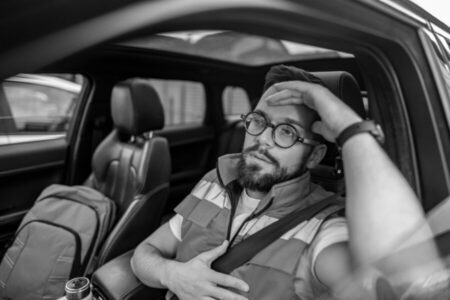 A bearded man wearing glasses and a vest sits in the driver's seat of a car, looking fatigued with one hand on his forehead and the other on his chest.