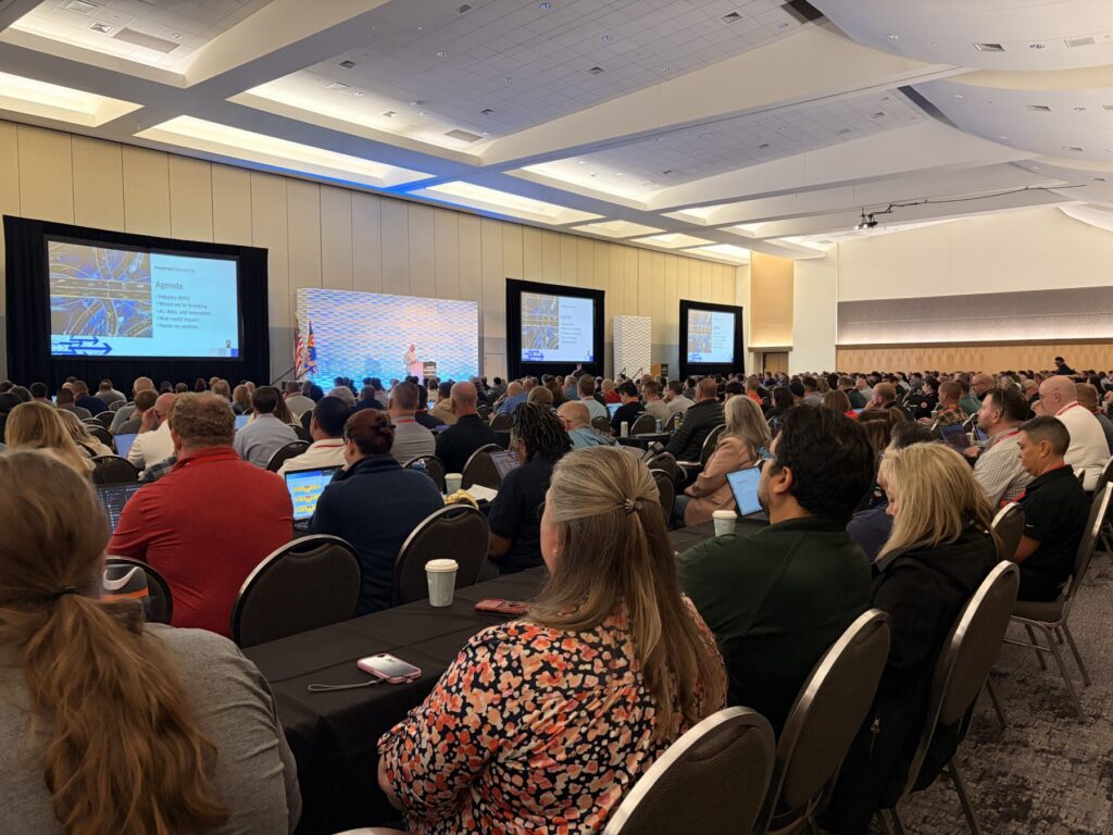 A large group of people seated in a conference room listening to a presenter on stage with slides projected on multiple screens.