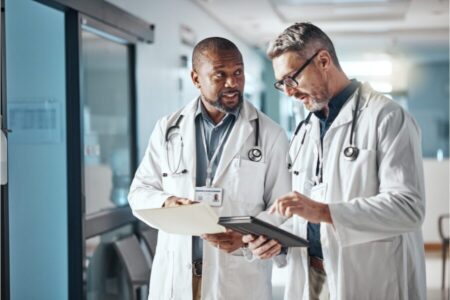 Two doctors in white coats stand in a hospital hallway, discussing patient information using a clipboard and a digital tablet.