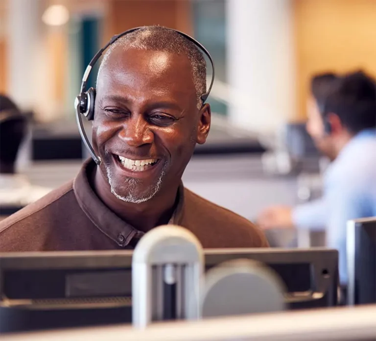 Smiling man wearing a headset sits at a desk in an office, looking at a computer screen. Another person is visible in the background.