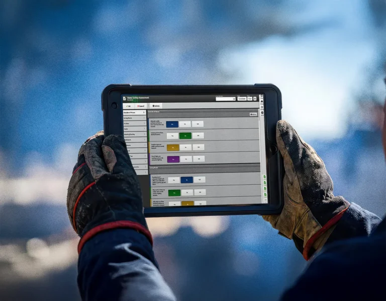Person wearing work gloves holds a tablet displaying a data management interface with various colored labels and status indicators, while outdoors.