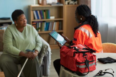 A healthcare worker in an orange uniform consults with an older woman holding a cane, with a red medical bag on a table in the foreground.