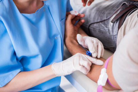 A healthcare worker in blue scrubs draws blood from a patient's arm using a syringe and tourniquet.
