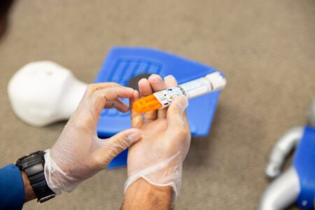 Gloved hands hold an epinephrine auto-injector next to blue medical training equipment on a carpeted surface.